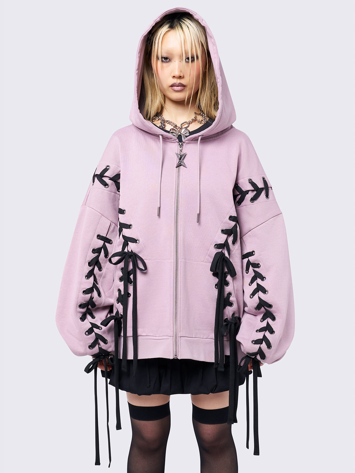 Pink zip-up hoodie with black lace corset details along the sleeves and front. Features a drawstring hood, zip fastening, long sleeves, and an oversized fit.