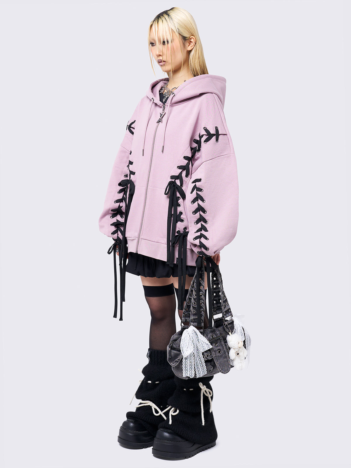 Pink zip-up hoodie with black lace corset details along the sleeves and front. Features a drawstring hood, zip fastening, long sleeves, and an oversized fit.