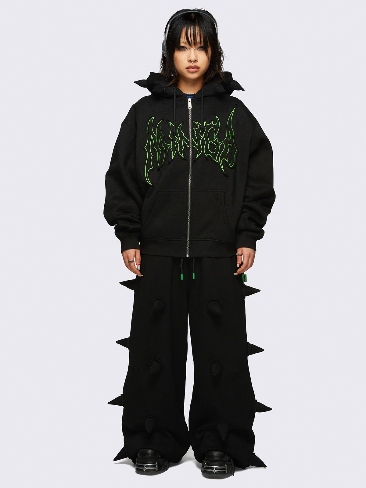 Black zip-up hoodie jacket featuring 3D spikes, green Minga embroidery, a drawstring hood, and zip fastening.