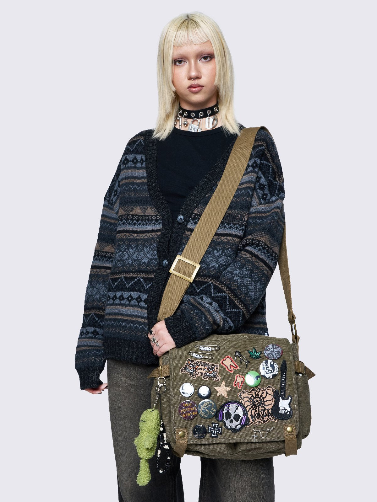 Olive green canvas messenger bag with grunge-inspired patches and pins, featuring a guitar, skull, alien, and other edgy designs. Includes a frog plush keychain and adjustable strap for a Y2K, alternative look.