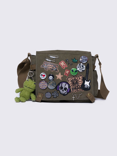 Olive green canvas messenger bag with grunge-inspired patches and pins, featuring a guitar, skull, alien, and other edgy designs. Includes a frog plush keychain and adjustable strap for a Y2K, alternative look.