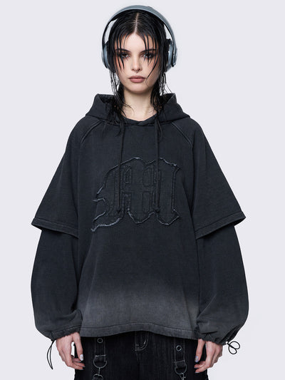 Oversized layered hoodie in washed black, featuring an embroidered "M" patch on the front, faded cuffs and hem, a drawstring hood, toggle elastic drawstrings, and raglan long sleeves.