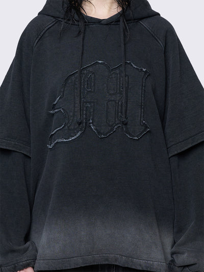 Oversized layered hoodie in washed black, featuring an embroidered "M" patch on the front, faded cuffs and hem, a drawstring hood, toggle elastic drawstrings, and raglan long sleeves.